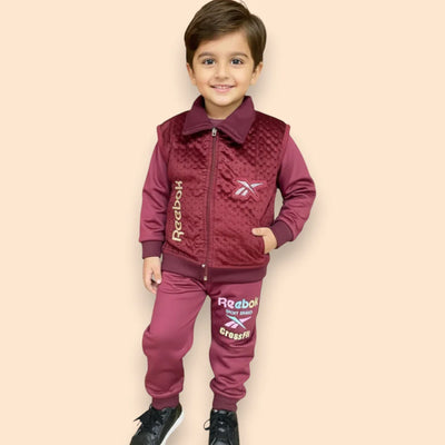 A young boy smiles while wearing a maroon Reebok tracksuit and black sneakers, standing against a plain beige background.