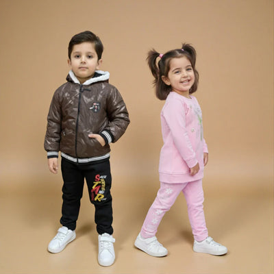 A young boy in a brown jacket and black pants stands next to a young girl in a pink outfit with pigtails. Both children are smiling and posing against a plain beige background.