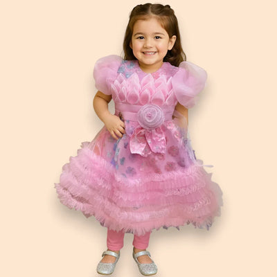 A young girl smiles while wearing a frilly pink dress with floral details, puffed sleeves, and a large flower on the waist, paired with pink leggings and silver shoes, standing against a beige background.